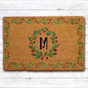 May include: A rectangular doormat with a brown coir surface. The mat features a green wreath with the letter "M" in the center, surrounded by red berries. A border of green leaves and red berries frames the mat.