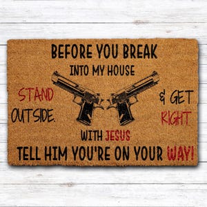 May include: A rectangular doormat made of natural fibers. The mat features two black gun illustrations and text in black and red, including "BEFORE YOU BREAK INTO MY HOUSE" and "TELL HIM YOU'RE ON YOUR WAY!"