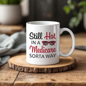 May include: A white ceramic mug featuring the black and red text Still Hot IN A Medicare SORTA WAY with a graphic of red cat-eye sunglasses positioned between the words Hot and Medicare on the front of the mug.