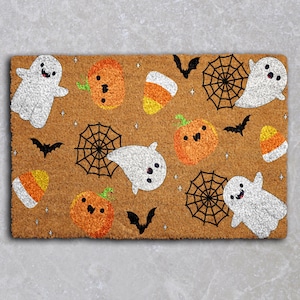 May include: A rectangular doormat with a brown background features Halloween-themed illustrations. White ghosts, orange pumpkins, candy corn, black spiderwebs, and bats are scattered across the mat. The design includes small star accents.