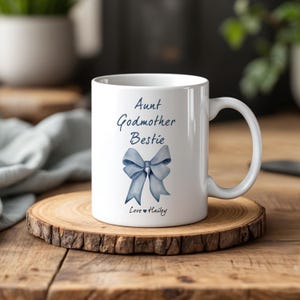 May include: A white ceramic mug featuring the dark blue text "Aunt Godmother Bestie" above a watercolor-style light blue bow illustration, with the text "Love ♥ Hailey" printed in dark blue at the bottom of the mug, centered below the bow.