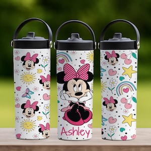 Op de afbeelding: Drie witte waterflessen met zwarte deksels en handvatten. Elke fles heeft een Minnie Mouse-ontwerp met roze strikken, harten, sterren en regenbogen. De middelste fles heeft de naam "Ashley" in roze.