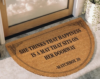 May include: A half-circle doormat made of natural fibers. The mat features the text "SHE THINKS THAT HAPPINESS IS A MAT THAT SITS ON HER DOORWAY -MATCHBOX 20" in black. Decorative border. Placed in front of a door.