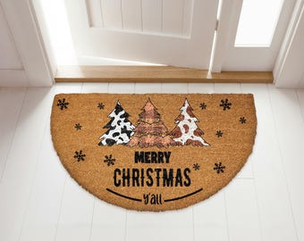 May include: A half-circle doormat made of natural fibers. It features three Christmas trees with cow print patterns and string lights. The text "MERRY CHRISTMAS y'all" is printed in black.