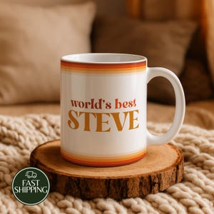 May include: A white ceramic mug featuring the text "world's best STEVE" in orange and brown lettering. The mug is decorated with horizontal stripes in shades of orange, yellow, and brown at the top and bottom edges, and has a curved handle.