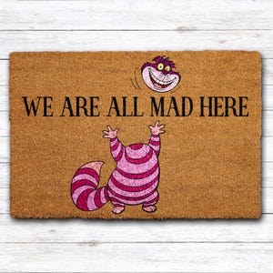 May include: A rectangular doormat with the text "WE ARE ALL MAD HERE" in black. The mat features a cartoon illustration of the Cheshire Cat from Alice in Wonderland, with a pink and white striped body and a smiling face.
