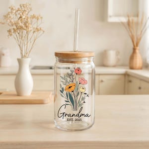 May include: A clear glass tumbler with a wooden lid and a glass straw. The tumbler features a floral design with orange, pink, and yellow flowers, and the word "Grandma" with "Est. 2025" below it. The tumbler is on a light-colored wooden surface.