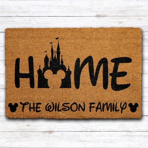 May include: A rectangular doormat made of natural coir fiber. The mat features the word "HOME" in large black letters, with a silhouette of a castle and Mickey Mouse ears. Below, the text "THE WILSON FAMILY" is displayed.