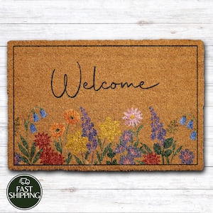 May include: A rectangular doormat made of natural coir fiber. The mat features the word "Welcome" in a handwritten style, above a colorful floral border with blue, orange, yellow, and purple flowers. A black border frames the mat.