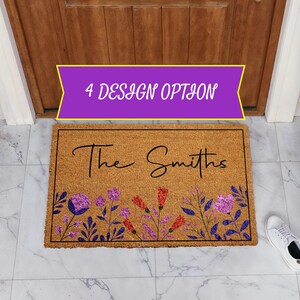 May include: A rectangular tan coir doormat featuring a black border and the text "The Smiths" in a cursive script. The bottom section displays a colorful floral pattern with purple, red, and blue flowers and leaves on a natural fiber surface.