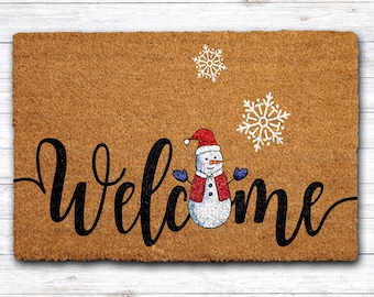 May include: A rectangular doormat with a brown coir surface. The word "Welcome" is written in black cursive script. A snowman wearing a red hat, vest, and blue mittens is in the center. Two white snowflakes are on the top right.