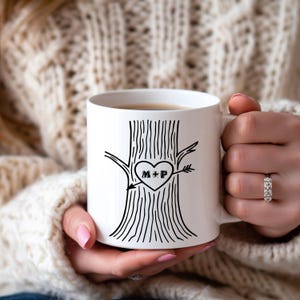 Peut inclure: Mug en céramique blanche avec un motif de tronc d'arbre noir et une sculpture en forme de cœur avec les initiales "M+P". La tasse est tenue par une personne portant un pull crème. La tasse est remplie d'un liquide foncé.
