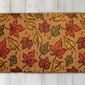 May include: A rectangular doormat with a repeating pattern of autumn leaves in shades of red, orange, yellow, and green on a brown background. The leaves are stylized with dark brown outlines, creating a seasonal, welcoming design.