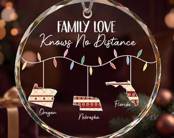 May include: A circular clear glass ornament featuring the text "FAMILY LOVE Knows No Distance" above a string of colorful Christmas lights. Three state shapes, Oregon, Nebraska, and Florida, hang from the lights, each with a red and white pattern.