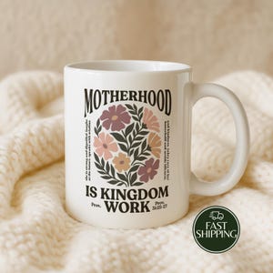 May include: A white ceramic mug featuring a floral graphic in muted tones of pink, orange, and brown. The text reads MOTHERHOOD IS KINGDOM WORK, along with the verse Prov. 31:25-27 and the phrase she is strong and dignified, laughs at the future, speaks with wisdom.