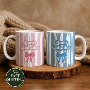 May include: Two ceramic mugs featuring vertical wavy stripes. The left mug is pink with a pink bow and the text 'Liam's Godmother'. The right mug is blue with a blue bow and the text 'Liam's Godfather'. Both mugs have white handles and interiors.