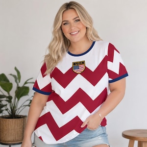 May include: White t-shirt with a red and white zig-zag pattern. The shirt has navy blue trim on the sleeves and neckline. A patch with the text "USA" and an American flag is on the chest.