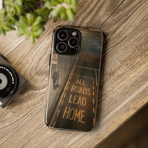 Puede incluir: Una funda de teléfono con una escena callejera con el texto "ALL ROADS LEAD HOME" en amarillo. La funda tiene un acabado brillante y una imagen detallada de una persona caminando por una acera. La funda está sobre una superficie de madera.