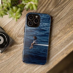 May include: A smartphone case featuring a graphic print of a person sitting on a diving board over deep blue ocean water. The case has a cutout for the camera lens array on the back and covers the rear and sides of the mobile device.
