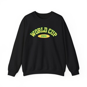 以下が含まれることがあります： 黒色のクルーネックのスウェットシャツ。緑と黄色の文字で「WORLD CUP 2026」と書かれています。テキストは、中央に2026年と書かれた黄色の楕円の上にアーチ状に配置されています。スウェットシャツは長袖で、リブ編みのネックラインです。