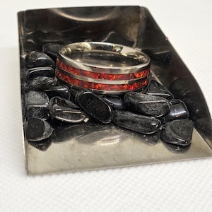 May include: A silver ring with two red bands of inlaid opal. The ring is sitting on a silver tray with black stones.