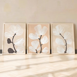 May include: Three framed botanical art prints. Each print features a white floral design with black stems and leaves against a beige background. The frames are a light wood color, and the overall aesthetic is minimalist and modern.