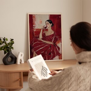 Op de afbeelding: Een ingelijste kunstdruk met een vrouw in een rode jurk die wijn drinkt. Het kunstwerk heeft een gedurfde, abstracte stijl met rode en witte tinten. Een boek met het woord "MATISSE" is zichtbaar.