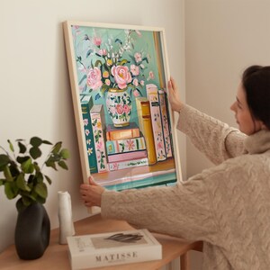 K&ouml;nnte beinhalten: Ein gerahmtes Gem&auml;lde mit einem Stillleben mit einer Vase voller rosa Blumen und einem Stapel B&uuml;cher. Die Vase ist mit Blumenmustern verziert und die B&uuml;cher haben bunte R&uuml;cken.