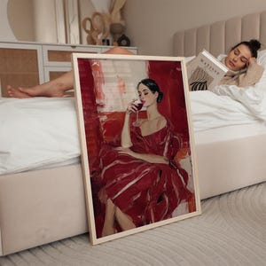 Op de afbeelding: Een ingelijste kunstdruk toont een vrouw in een rode jurk die wijn drinkt. Het kunstwerk heeft een rood-wit kleurenschema, waarbij het donkere haar en de huid van de vrouw een contrast vormen. De print wordt op een bed getoond, met een lezende persoon op de achtergrond.