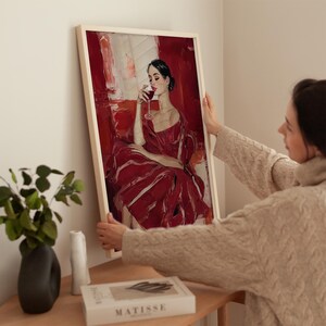 Op de afbeelding: Ingelijste kunstdruk met een vrouw in een rode jurk die wijn drinkt. Het kunstwerk heeft een textuurrijke, schilderachtige stijl met een rood en wit kleurenpalet. De lijst is licht van kleur. De afdruk wordt door een persoon vastgehouden.