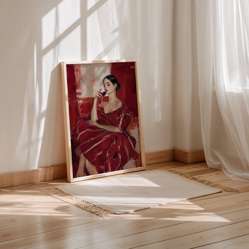 Op de afbeelding: Een ingelijste kunstdruk toont een vrouw in een rode jurk, die een glas wijn vasthoudt. Het kunstwerk heeft een rood-wit kleurenschema, met een houten lijst. De print leunt tegen een witte muur, met een vloerkleed op de houten vloer.