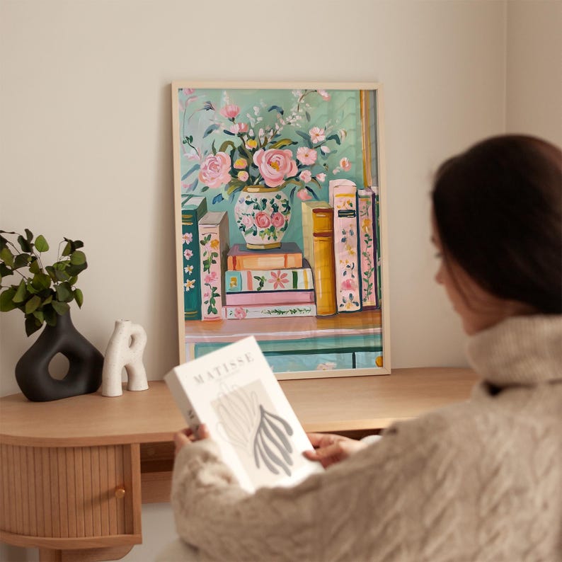 K&ouml;nnte beinhalten: Ein gerahmtes Gem&auml;lde mit einer Vase voller rosa Blumen mit gr&uuml;nen Bl&auml;ttern, die auf einem Regal mit einem Stapel B&uuml;cher steht. Die B&uuml;cher haben bunte Umschl&auml;ge mit Blumenmustern. Das Gem&auml;lde hat einen hellgr&uuml;nen Hintergrund.