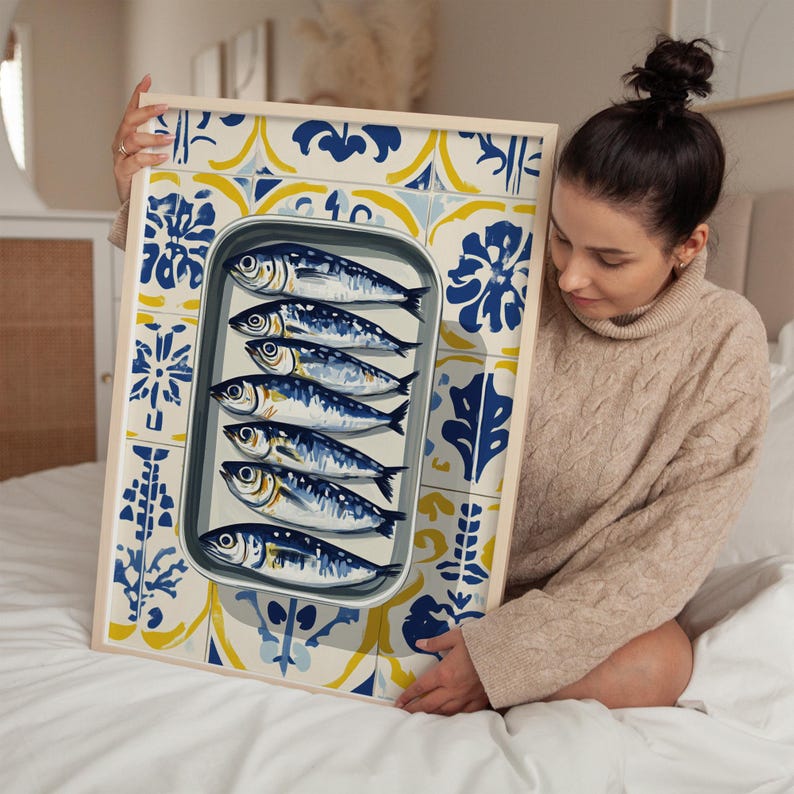 May include: A framed artwork featuring a tin of sardines. The sardines are painted with blue and silver details. The frame has a decorative tile design in blue, yellow, and white. The artwork is held up by a person.