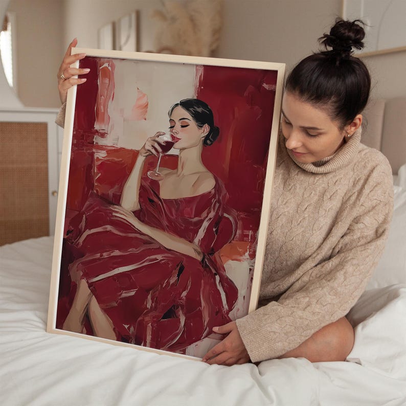 Op de afbeelding: Een ingelijste kunstdruk met een vrouw in een rode jurk, genietend van een glas rode wijn. Het kunstwerk heeft een gedurfde, schilderachtige stijl met een overwegend rood kleurenpalet. De lijst heeft een lichte houtkleur.