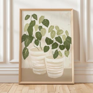 Może przedstawiać: Oprawiony wydruk artystyczny z ilustracją botaniczną. Dwie rośliny doniczkowe z dużymi zielonymi liśćmi przedstawione są w białych i beżowych donicach w paski. Grafika ma neutralną paletę kolorów z jasnobeżowym tłem i ramą z naturalnego drewna.