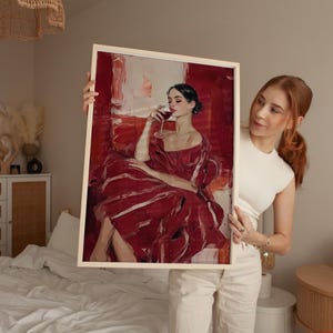 Op de afbeelding: Een ingelijste kunstdruk met een vrouw in een rode jurk, zittend en een glas wijn vasthoudend. Het kunstwerk heeft een gedurfde, schilderachtige stijl met rode en witte tinten. Het frame is licht van kleur.
