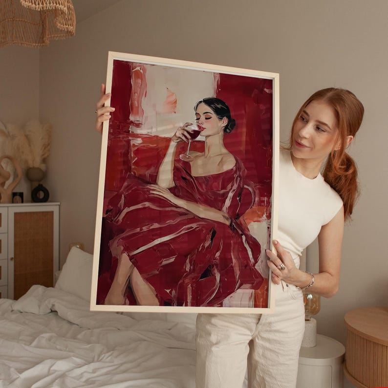 Op de afbeelding: Een ingelijste kunstdruk met een vrouw in een rode jurk, zittend en een glas wijn vasthoudend. Het kunstwerk heeft een gedurfde, schilderachtige stijl met rode en witte tinten. Het frame is licht van kleur.