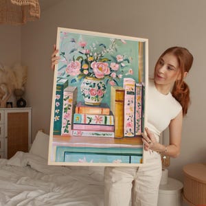 K&ouml;nnte beinhalten: Ein gerahmtes Gem&auml;lde mit einem Blumenarrangement in einer blau-wei&szlig;en Vase und einem Stapel B&uuml;cher dahinter. Die Blumen sind rosa, wei&szlig; und gr&uuml;n, die B&uuml;cher sind in verschiedenen Braunt&ouml;nen und Rosa gehalten.
