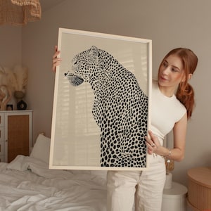 May include: A framed artwork featuring a black and white leopard illustration. The leopard is depicted in profile with a spotted coat. The artwork is encased in a light-colored wooden frame, suitable for home decor. The background is a neutral beige.