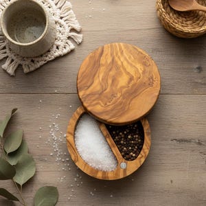 May include: A wooden salt and pepper cellar with a lid, filled with white salt and black peppercorns. The container is made of olive wood and sits on a wooden surface. A small woven basket with a wooden spoon and a ceramic mug are also visible.