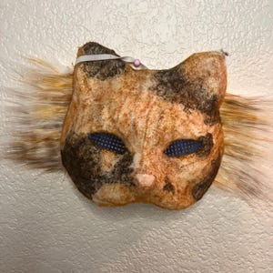 May include: A handmade calico cat mask with a furry orange and white tail. The mask has blue mesh eyes and a pink nose.