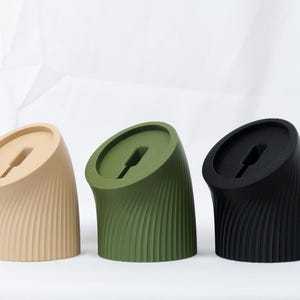 May include: Three phone stands in different colors: beige, green, and black. Each stand has a curved, ribbed design and a slot for charging.