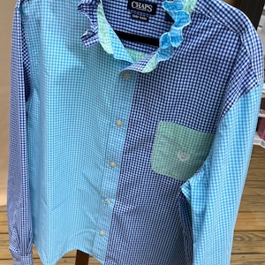 May include: A long-sleeved button-down shirt with a patchwork design in shades of blue and white gingham. The shirt features a ruffled collar in blue and green, a chest pocket, and the "CHAPS" logo.