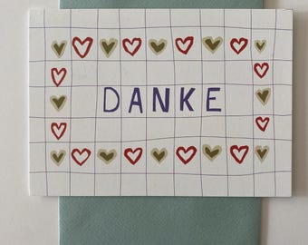 May include: A white card with a grid pattern and the word "DANKE" in purple. The card is decorated with red and gold hearts. It rests on a teal envelope, creating a simple, yet elegant design.