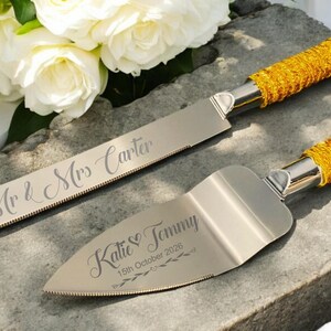 May include: A silver wedding cake knife and server set with gold handles. The knife is engraved with "Mr & Mrs Carter", and the server with "Katie & Tommy 15th October 2026". White flowers are in the background.