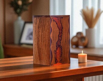 May include: A handcrafted wooden box with a vibrant red resin river running through the center. The box is made of dark brown wood and sits on a wooden table. A lit candle is next to the box.
