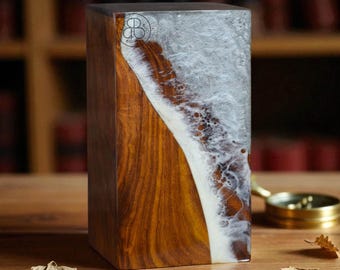 May include: A rectangular wooden block with a unique design. The block features a combination of dark brown wood and a clear resin with a white, foamy pattern. A small logo is visible on the top. The item is displayed on a wooden surface.