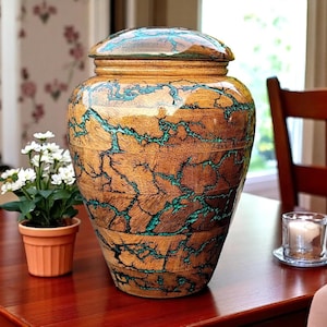 May include: A large, wooden urn with a lid, featuring a unique design of brown wood with turquoise-colored accents. The urn is on a dark wooden table, with a small potted plant and a candle nearby. The urn is a decorative item.
