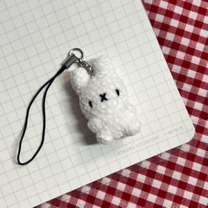 May include: A white plush bunny charm with a black cross for a mouth and black eyes. The bunny is attached to a silver-colored metal ring and a black cord. The charm is on a white grid paper.
