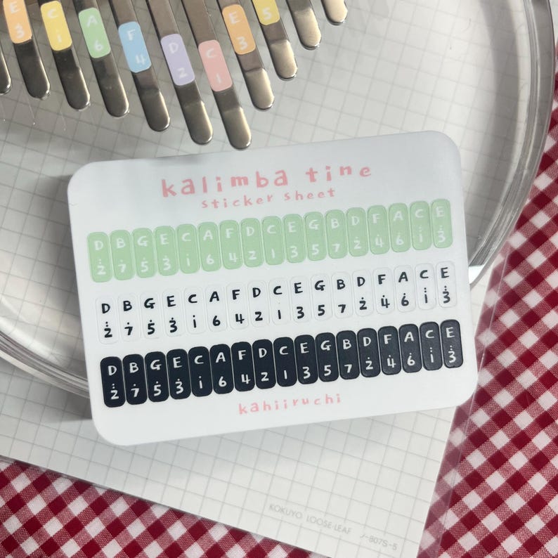 Pu&ograve; includere: Un foglio di adesivi per kalimba con etichette verdi e nere con note musicali e numeri. Il testo "kalimba tine sticker sheet" &egrave; in rosa. Il marchio "kahiiruchi" &egrave; in basso.